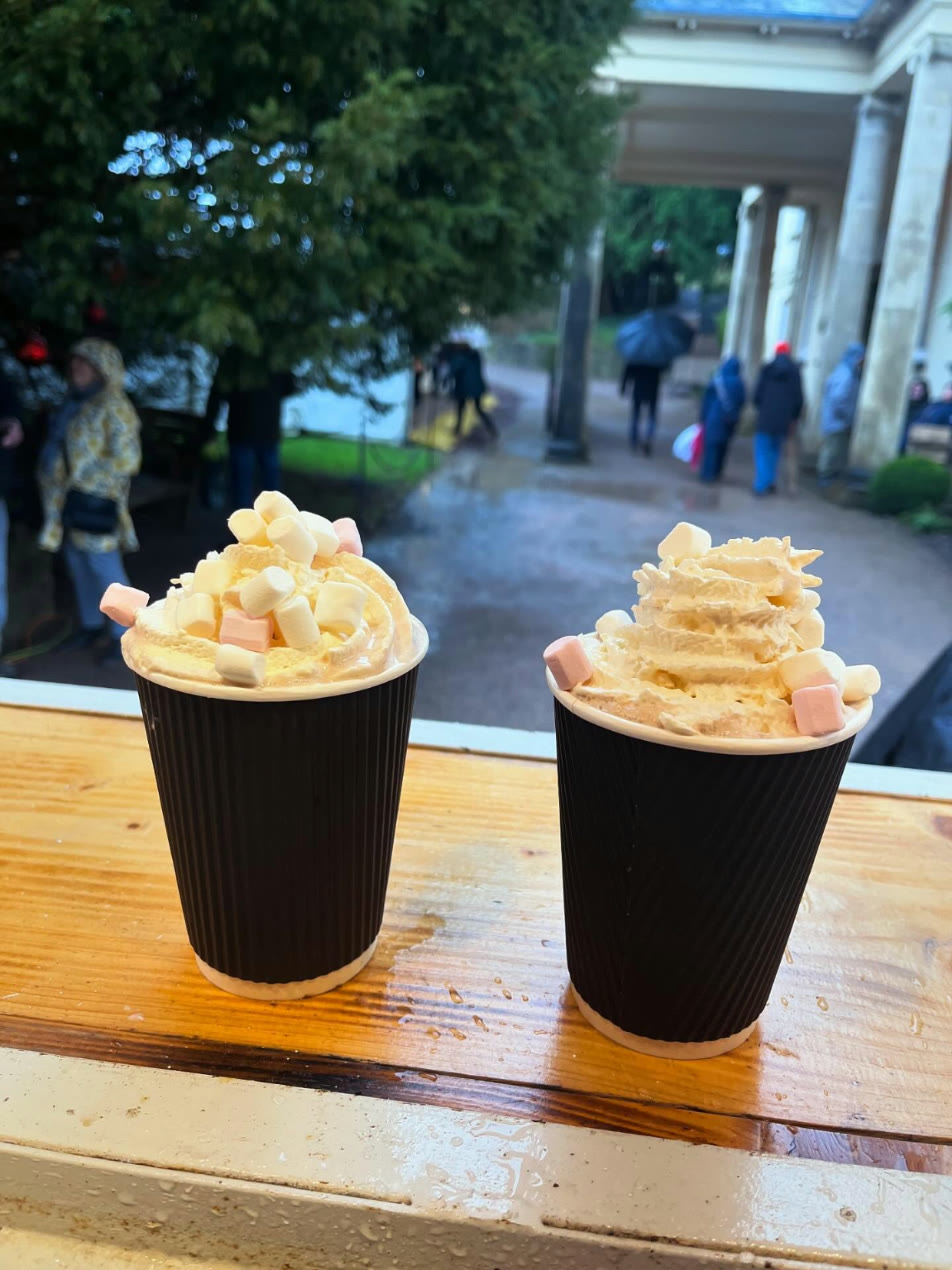 Two hot chocolates topped with cream and marshmallows on the horsebox bar, Aberglasney Gardens in the rain behind