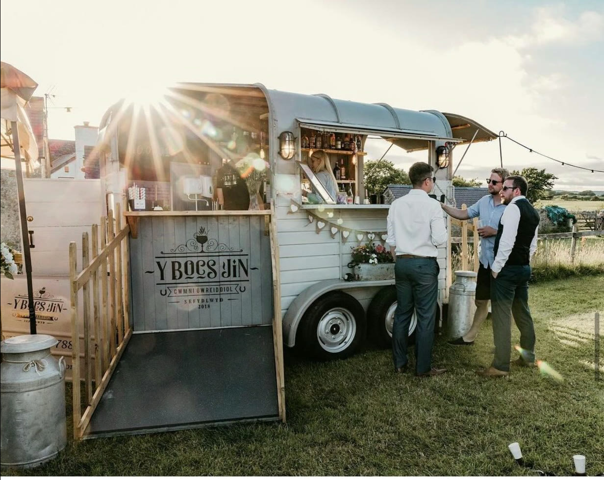 The Y Bocs Jin horsebox bar at a summer wedding, guests at the hatch, sun low behind the trailer